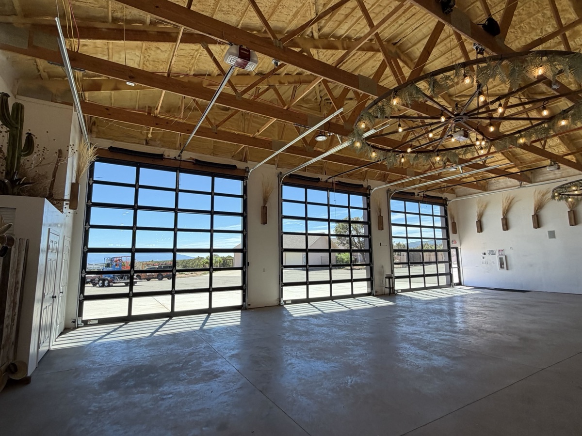 Interior of a commercial event space with three full-view glass garage doors, wood-truss ceiling, and string lights