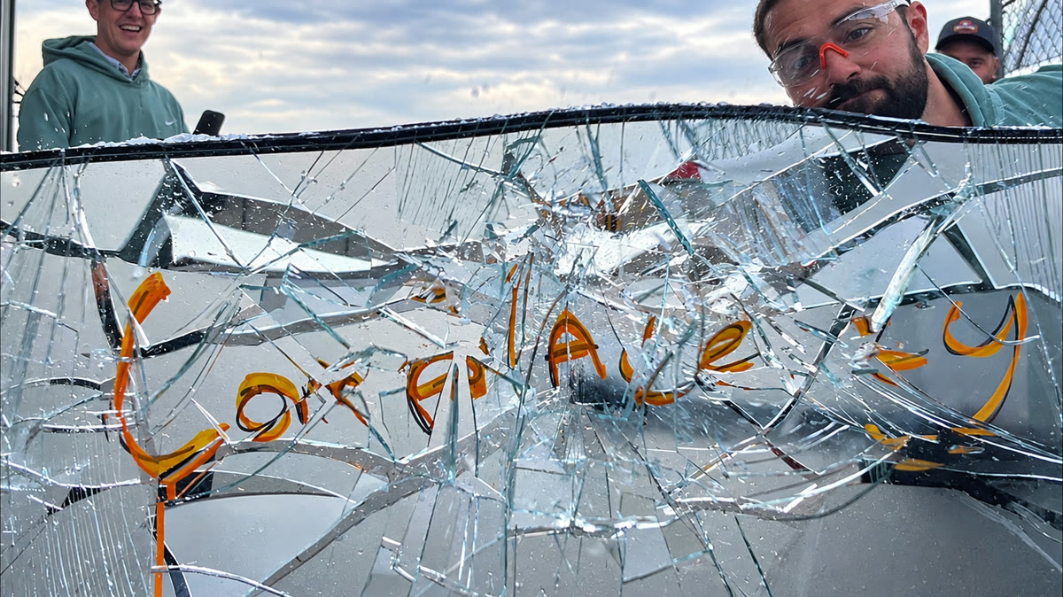 A pane of glass smashed into a spiderweb of cracks, a word written across it in orange marker now broken apart, three men in Kooler green looking on from behind it