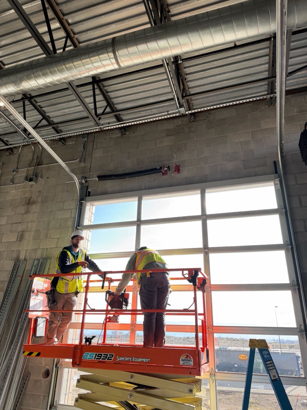 Two Kooler Kraftsmen on a scissor lift installing a full-view commercial garage door in a block building.