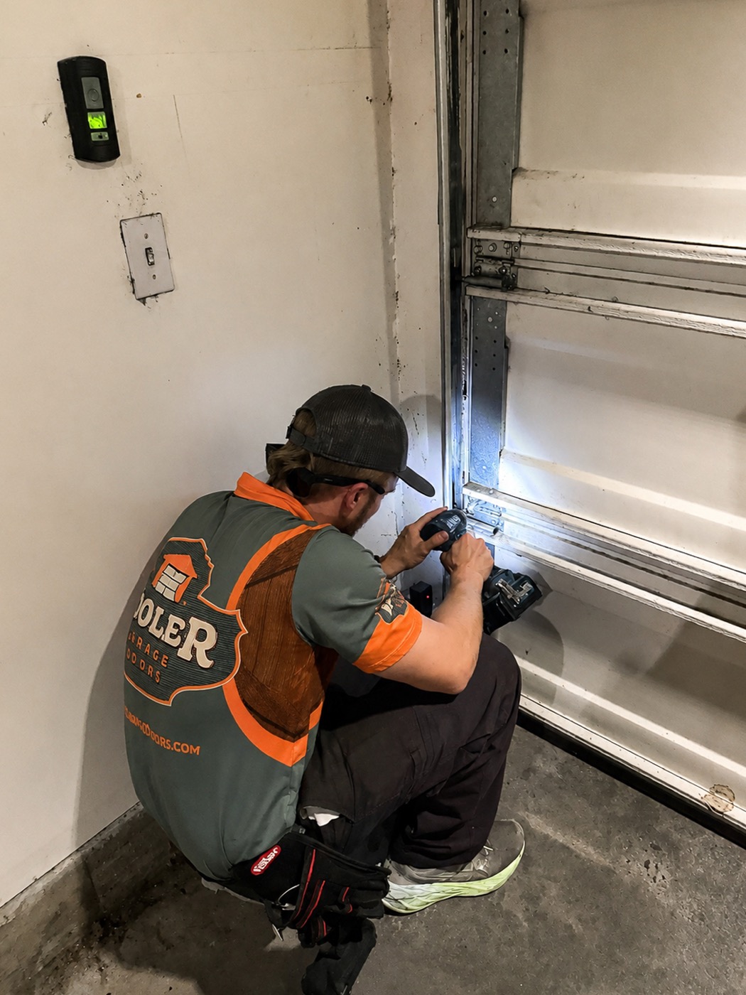 A Kooler Kraftsman crouched at the bottom of a garage door track with a drill.