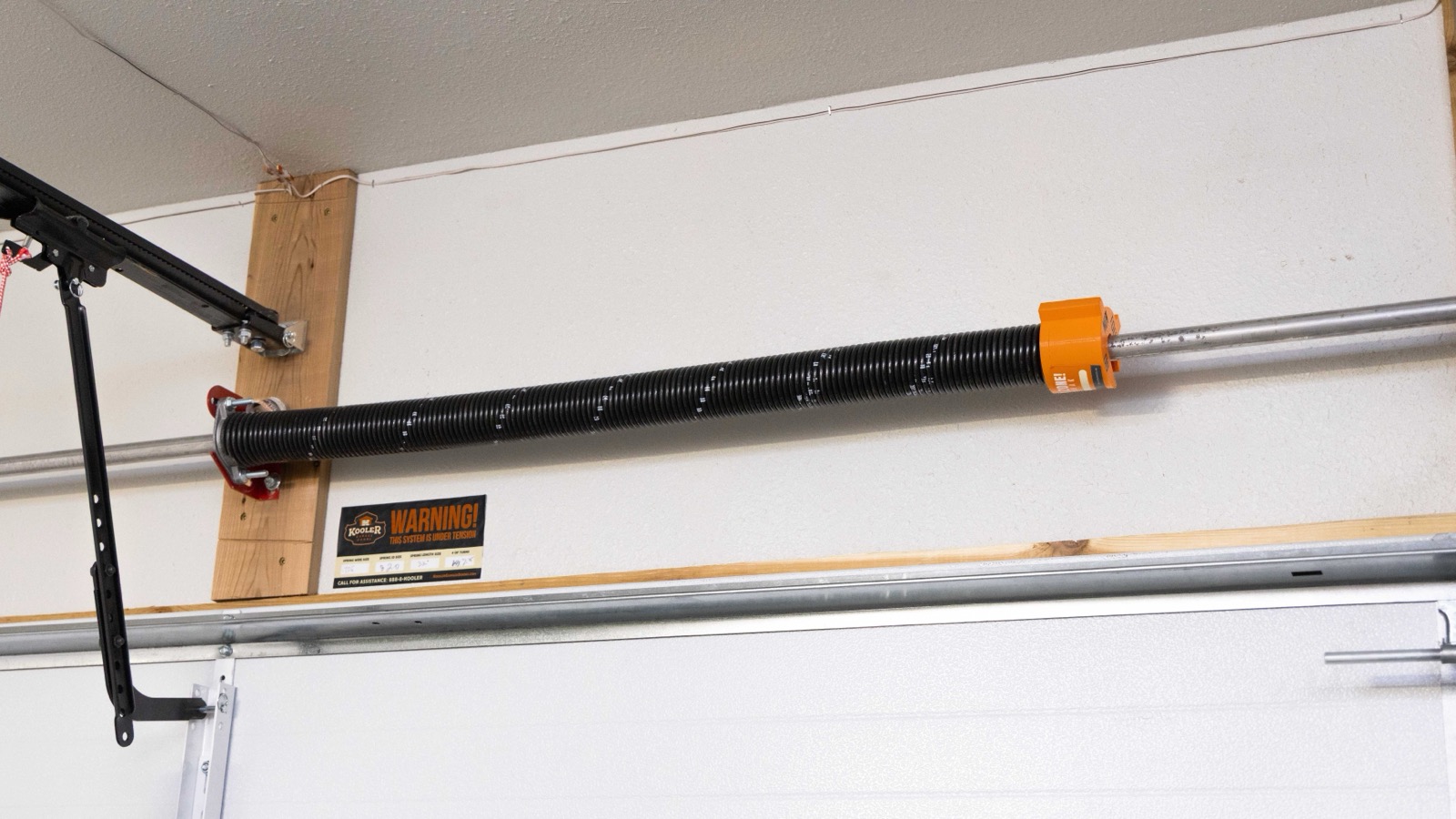 A torsion spring mounted above a garage door with an orange K Kap safety warning cap on the winding-cone end of the shaft