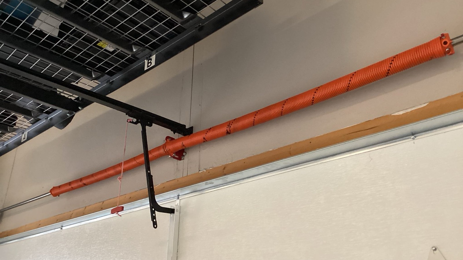 A torsion spring above a closed garage door, orange steel wound along a shaft, its black center bracket bolted to the wall